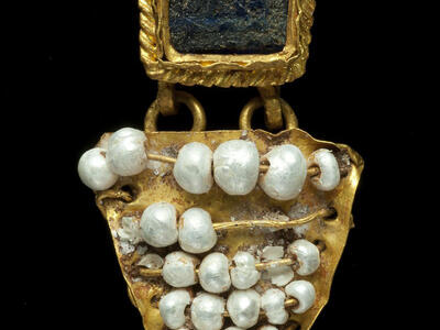 A gold earring with a square blue or black stone at the top, bordered by twisted gold, and a triangular gold section below, decorated with irregular white mother-of-pearl beads arranged in rows.