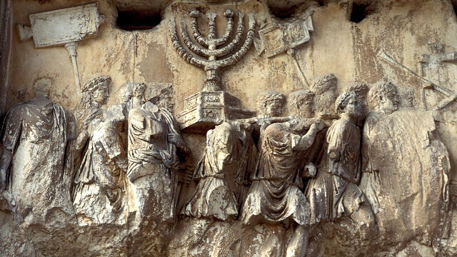 Ancient, weathered stone relief showing people in robes carrying a large menorah and other objects in a procession.