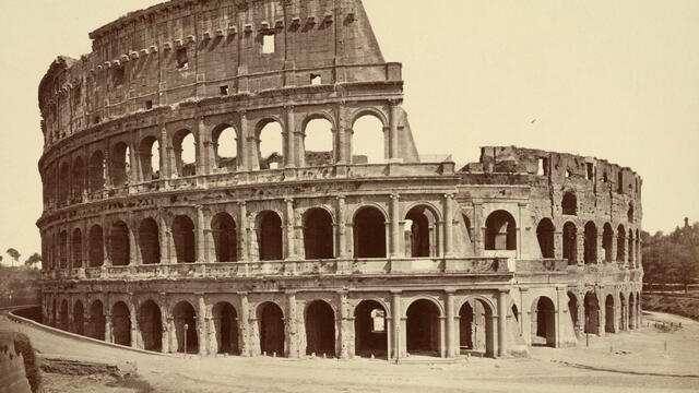 Black-and-white photo of a large amphitheater, showing its ancient stone arches and partial ruin. The large amphitheater stands alone, with some missing sections and an empty foreground.