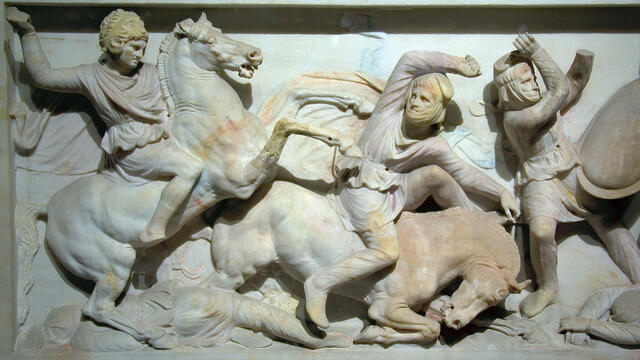 A marble relief sculpture shows three figures in battle: a mounted warrior attacks another figure who is falling from a horse, while a third figure takes a defensive pose. Two additional figures, apparently dead, lie on the ground.