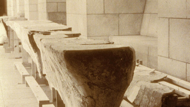 Wooden supports in the shape of a long, narrow table and bench with slabs of broken plaster on top, in a room with arched ceilings and stone walls.