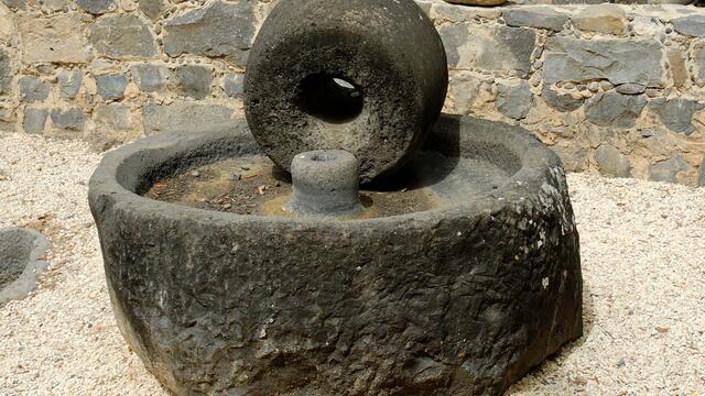 A large, round olive press consisting of a circular basin and wheel sits outdoors on gravel, surrounded by a low stone wall and other stone objects.