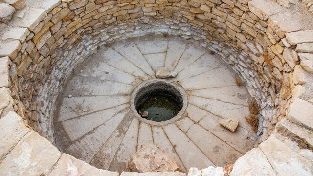 Circular stone basin with layered stone walls, a base with a radial pattern, and a round opening at the bottom. A few rocks are scattered inside.