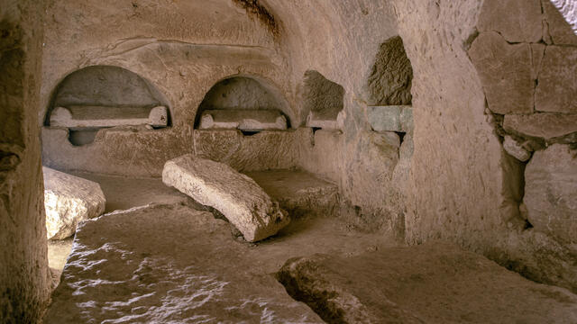 Ancient stone chamber with arched alcoves carved into the walls and rectangular stone slabs on the ground.