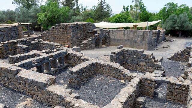 Stone ruins with partial walls, including some with window-like openings, surrounded by trees and greenery; shade cloths are stretched above part of the site.