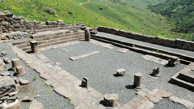Ruins of an ancient stone structure sit on a gravel surface, surrounded by low stone walls and scattered columns, overlooking a lush, hilly landscape.