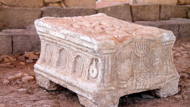 A carved stone object with detailed reliefs, including menorahs and arches, resting on four short legs, sits on a dirt floor among weathered stone blocks and ruins.