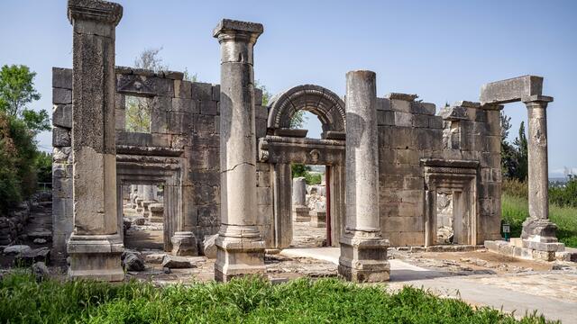 Partially preserved stone building facade with an arched entrance and four columns in front. Other stone ruins are visible through the entryway.