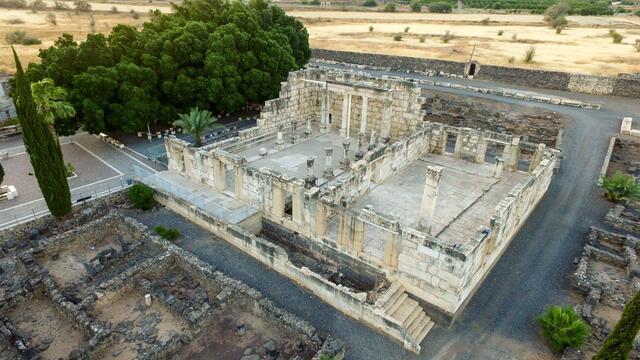 Aerial view of an ancient stone synagogue ruin surrounded by greenery and dry fields, with remnants of walls and columns visible, located near a tree-lined area and dirt pathways.