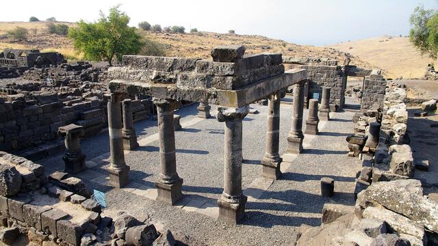 Ancient stone ruins of a rectangular structure with several columns and partial walls, set in a dry, hilly landscape with sparse vegetation under a clear sky.