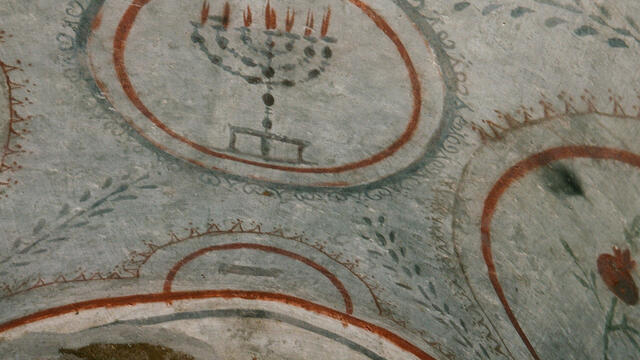 Ancient painting featuring a menorah with seven lit candles inside a bordered circle, surrounded by decorative patterns and arches on a stone surface.