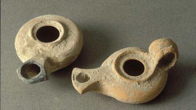 Two ancient, round clay oil lamps with spouts and central openings rest on a plain surface. The lamps are worn and show earthy tones.