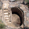 A round, stone-lined pit with built-in stone steps leading down to a dirt floor. The structure is surrounded by rocks and sparse vegetation.