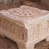 A carved stone object with ornate patterns and symbols, including rosettes or wheels within arches, resting on four short legs, sits on a dirt floor among weathered stone blocks and ruins.