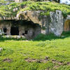 Ancient rock-cut tombs set into a grassy hillside, with arched entrances and stone walls partially covered by green vegetation. One entrance is barred with metal grates.