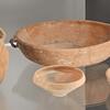 Four ancient, weathered ceramic vessels: a lidded jar with handles, a large bowl, a small bowl, and a shallow plate, displayed on a glass shelf against a plain, neutral background.