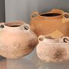 Three ancient clay pots with rounded bodies and small handles are displayed on a glass shelf. The pots vary in size and color, with some showing signs of wear and repair.