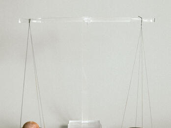 Balance Scale Reconstructed scale with two ancient bronze pans suspended from central beam of clear modern material. The left pan holds a balance weight and the right holds a piece of silver.