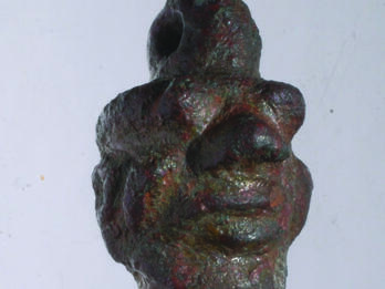 Bronze amulet of head with human and animal features and loop on top.