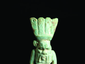 Frontal and back faience amulet of figure who is nude with feathered headdress and a grotesque face and beard.