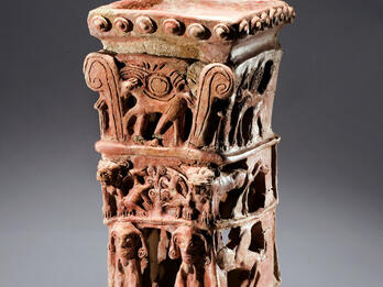Ceramic offering stand with flat top and four layers of decoration: bottom layer of nude female with raised arms flanked by lions, second layer of two winged figures, third layer of goats eating leaves off tree, and fourth layer of four-legged animal with winged sun disk and palm fronds. 