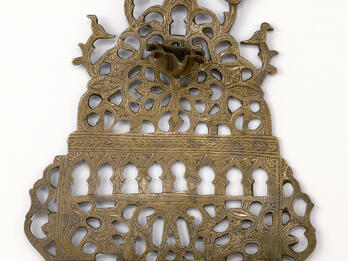 Hanukkah Lamp (Morocco) Oil lamp with cut-outs in various shapes, with row of eight cups on bottom and one cup on top.