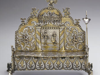 Hanukkah Lamp (Frankfurt am Main) Silver oil lamp with row of eight cups on bottom, and elaborate carvings of two figures and floral designs on lamp with carved birds on top.