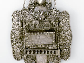 Silver Torah Shield Engraved shield of flowers, grapes, and other decorative elements around rectangular Hebrew inscription.