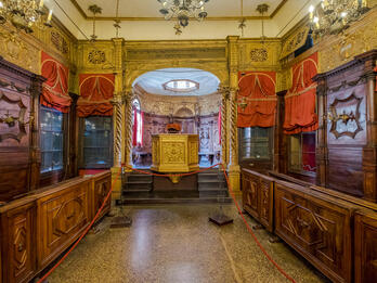Scuola Canton Synagogue, Venice Photograph of room lined with narrow wooden pews and set of stairs at far end of room leading to gilded podium.