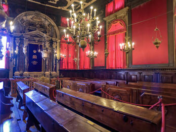 Scuola Levantina, Venice Photograph of room with rows of pews, curtains on right-hand wall, and columned archway on back wall.