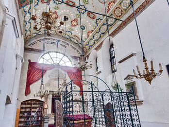 Rema Synagogue, Krakow Photograph of room with decorated ceiling, central platform with wrought-iron gate, and bookshelf on far wall.