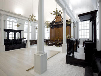 Tzedek ve-Shalom Synagogue, Suriname Photograph of rectangular room with arched windows, floor covered in sand, wooden cabinet on far wall, wooden benches on left side and along walls, and wooden lectern on right side.