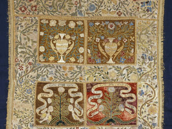 Cloth embroidered with floral border, and four central panels, the top two with pitchers, vines, and flowers, and the bottom two with trees and ribbons with Hebrew text. 