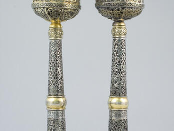 Two finials engraved with designs on the base and minaret-shaped tops with Hebrew text visible through cut-out windows. 