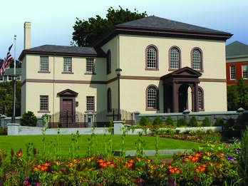 Touro Synaguge Photograph of exterior of building with semi-circular windows featuring American flag on left and flower garden in front.