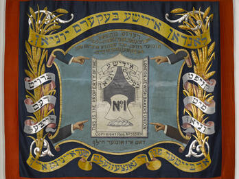 Banner of the London Bakers’ Union Banner with four arms pointed toward a crest in the center featuring a loaf of bread and English text, framed by Yiddish text and sheaves of wheat.