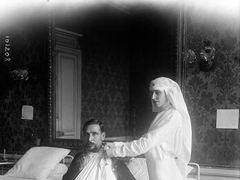 Ida Rubinstein as a Nurse Photograph of woman in veil standing next to bed where man sits with arm in sling.