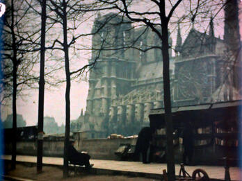 Book Seller and Notre Dame on the Quai de la Seine Photograph of a sidewalk and large cathedral looming behind it.