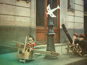 A "Taube" Is Spotted; A French 75 Is Immediately Put into Battery While "Pépéte," the Aviator, Prepares to Give Chase Photograph of a sidewalk where two boys crouch behind a play cannon and another boy sits in a handmade plane.