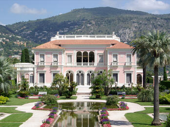 Villa Ephrussi de Rothschild Photograph of house surrounded by gardens and fountain and flanked by two tall palm trees, with mountains in background.
