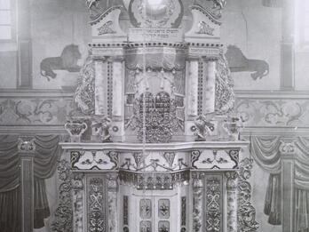 Pavoloch Torah Ark Photograph of large, ornate structure decorated with flourishes, lions, and vines, with three men in hats and beards standing in front of two closed doors at the bottom, and several other figures sitting in benches in foreground.
