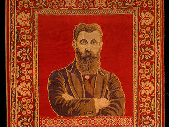Rug depicting a bearded man with crossed arms on top half, and two columns and hanging lamp on bottom half, surrounded by decorative border.