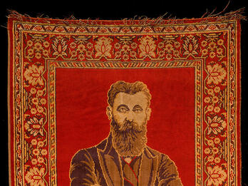 Rug depicting a bearded man with crossed arms on top half, and two columns and hanging lamp on bottom half, surrounded by decorative border.