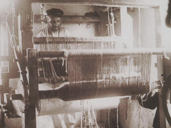 Tallis Weaver Photograph of large, multilayered loom with figure working on it.