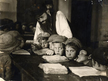 Teacher and Students in a Heder, Lublin Photograph of young boys sitting at a long table with books open as teacher leans over them to teach.