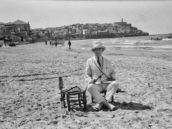 Photograph of man sitting on beach in hat and suit with hookah pipe in hand. 