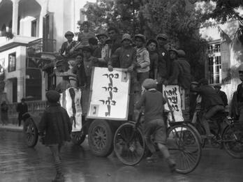 Photograph of men huddled together on a car holding billboards, surrounded by young men on bicycles following them down the street.