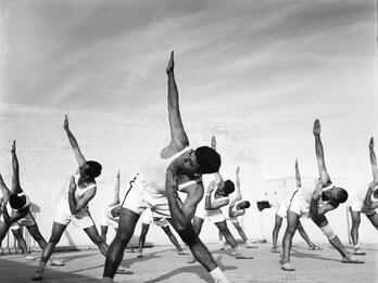 Sport Photograph of rows of men in athletic clothing bending at the waist with their right arms pointed up and their left arms bent in an angle toward their face.