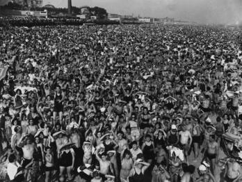 Aerial photograph of hundreds of people crowded together on beach with amusement park in background.