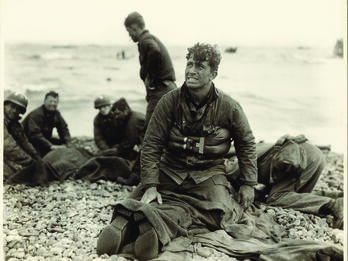 D Day Rescue, Omaha Beach, France. June 6, 1944 Photograph of soldiers on the beach, with man in center facing the viewer and looking up, as he kneels on top of a prone soldier.